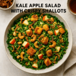 Overhead view of a kale apple salad in a ceramic bowl, topped with crispy shallots, golden fried cheese cubes, roasted cashews, and diced apples, styled on a light background with text overlay reading “Kale Apple Salad with Crispy Shallots."