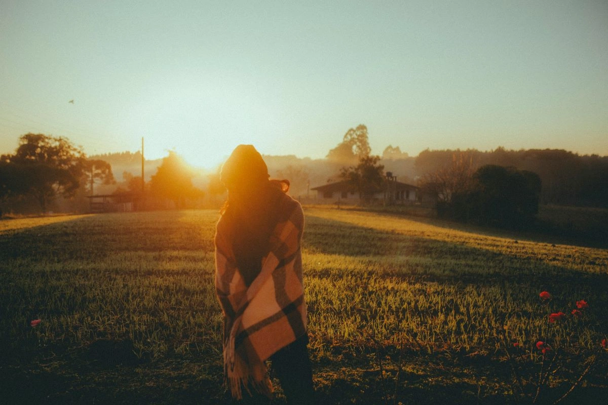 A woman standing in a field with the sun setting behind her, looking out at a homestead