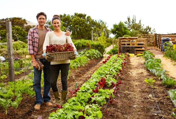 A woman in work boots and garden gloves, smiling in the dirt while holding freshly harvested vegetables.